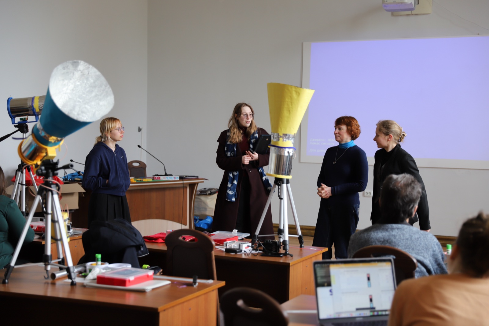 A presenter speaking next to a horn telescope in front of a projection screen during the workshop.